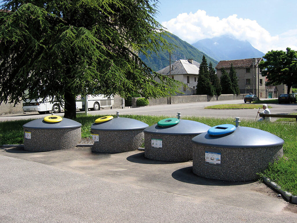 Vier Halbunterflur-Container mit kuppelförmigen, grauen Aufsätzen stehen nebeneinander am Rand eines asphaltierten Weges