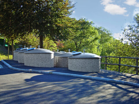 Vier Halbunterflur-Container mit kuppelförmigen, grauen Aufsätzen stehen nebeneinander am Rand eines asphaltierten Weges mit üppigem, sonnigem Grün im Hintergrund