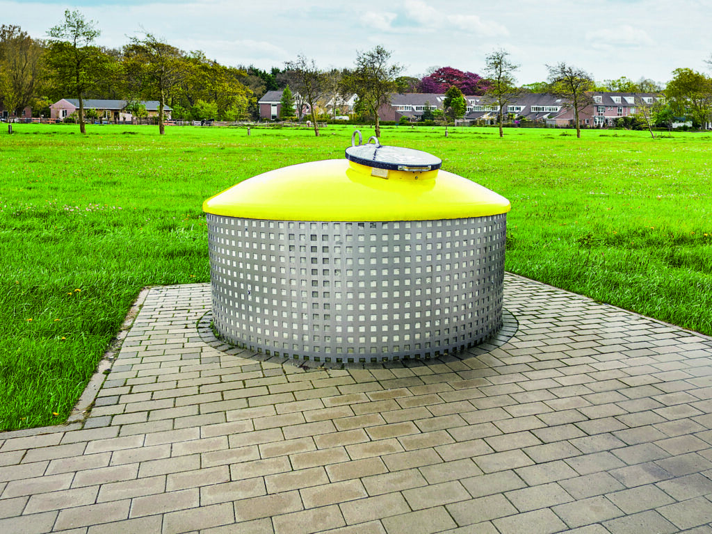 Ein Halbunterflur-Container mit kuppelförmigen, gelbem Aufsatz steht auf einer asphaltierten Fläche vor einer großen Wiese