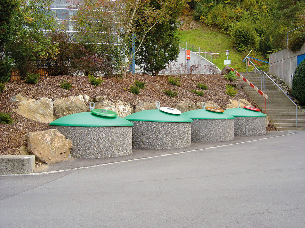 Vier Halbunterflur-Container mit kuppelförmigen, dunkelgrünen Aufsätzen stehen am Rand einer asphaltierten Straße