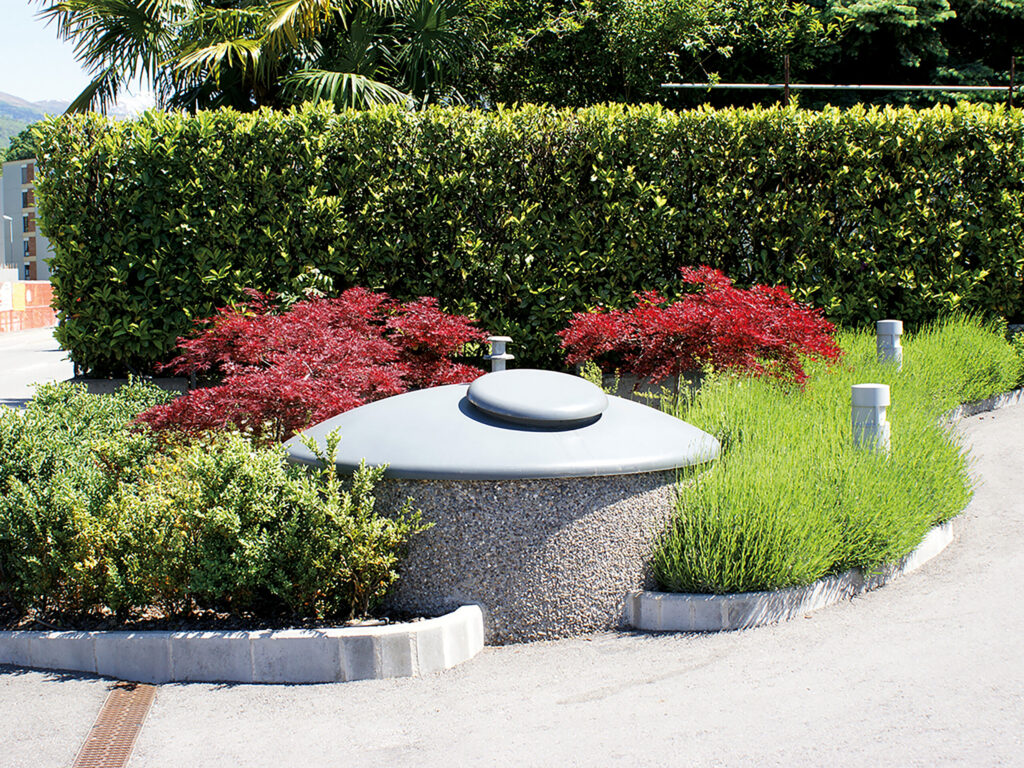 Ein Halbunterflur-Container Cupola mit kuppelförmigen, grauem Aufsatz steht am Rand eines asphaltierten Weges, umgeben von Sträuchern und dahinter eine grüne Hecke