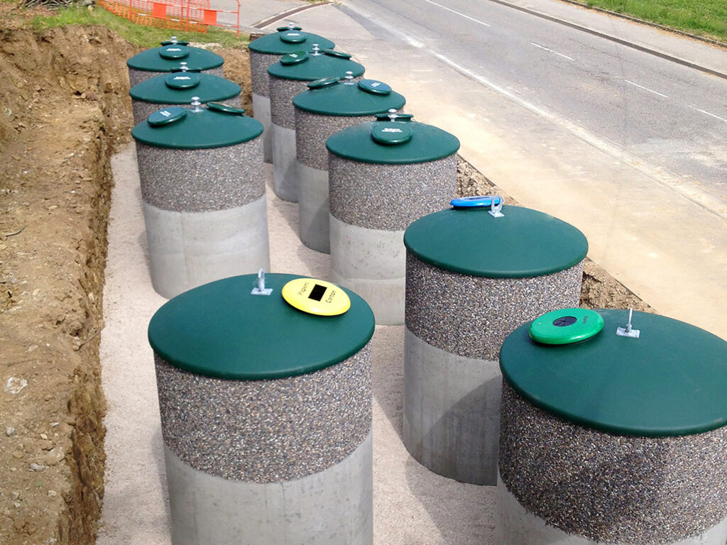 Mehrere Halbunterflur-Container mit kuppelförmigen, dunkelgrünen Aufsätzen stehen am Rand einer asphaltierten Straße