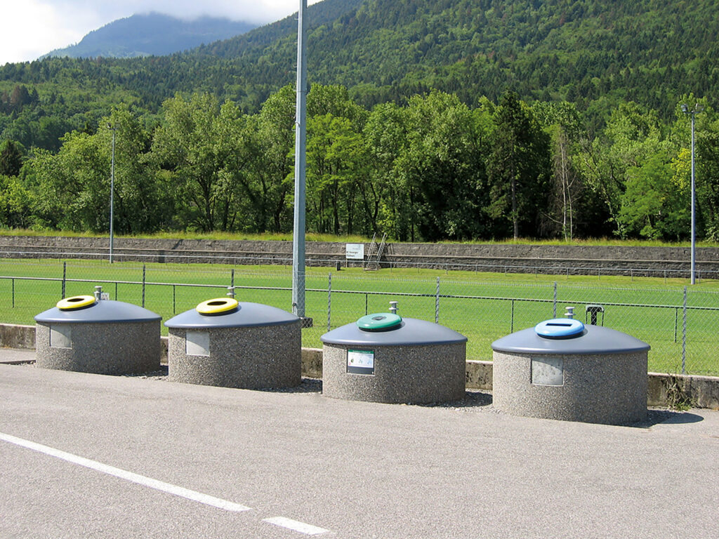 Vier Halbunterflur-Container mit kuppelförmigen, grauen Aufsätzen stehen nebeneinander am Rand eines asphaltierten Weges mit üppigem Grün im Hintergrund