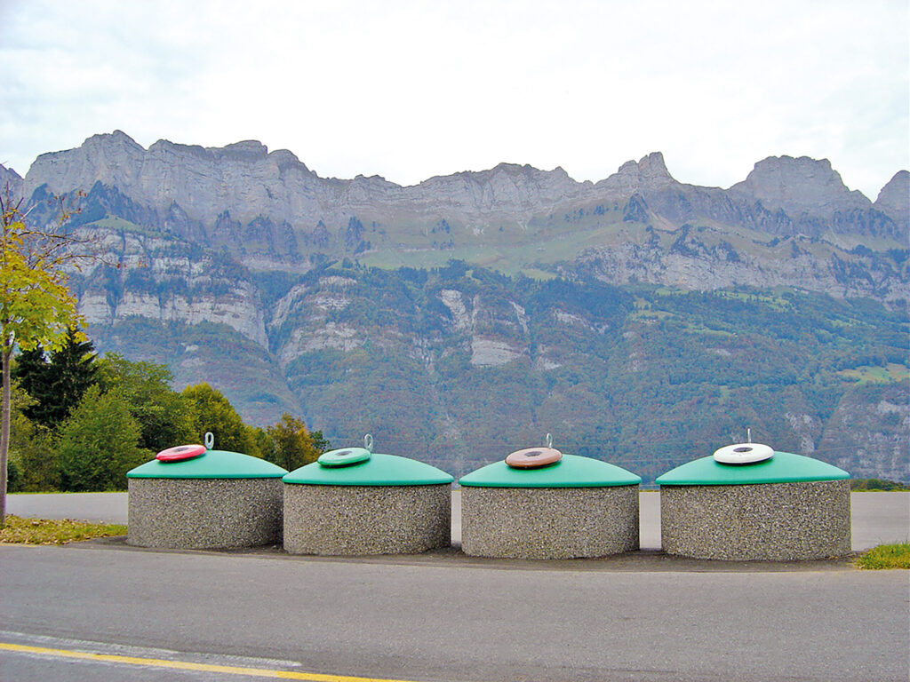 Mehrere Halbunterflur-Container mit kuppelförmigen, grünen Aufsätzen stehen nebeneinander am Rand einer Straße, Berge im Hintergrund