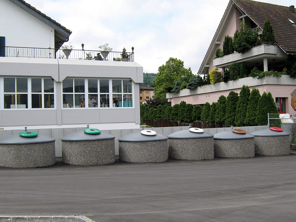 Mehrere Halbunterflur-Container mit kuppelförmigen, grauen Aufsätzen stehen nebeneinander an einem asphaltierten Platz vor einem Gebäude