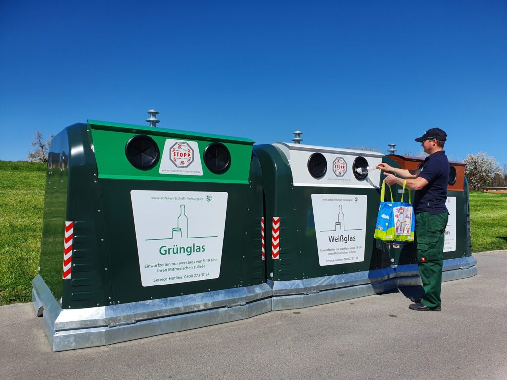 Zwei SMART-Line Glascontainer, einen für Bunt- und einen für Weißglas stehen an einem asphaltierten Platz. Eine Person wirft Gegenstand in die Einwurfsäule eines SMART-Line-Glascontainers. – Villiger Entsorgungssysteme AG